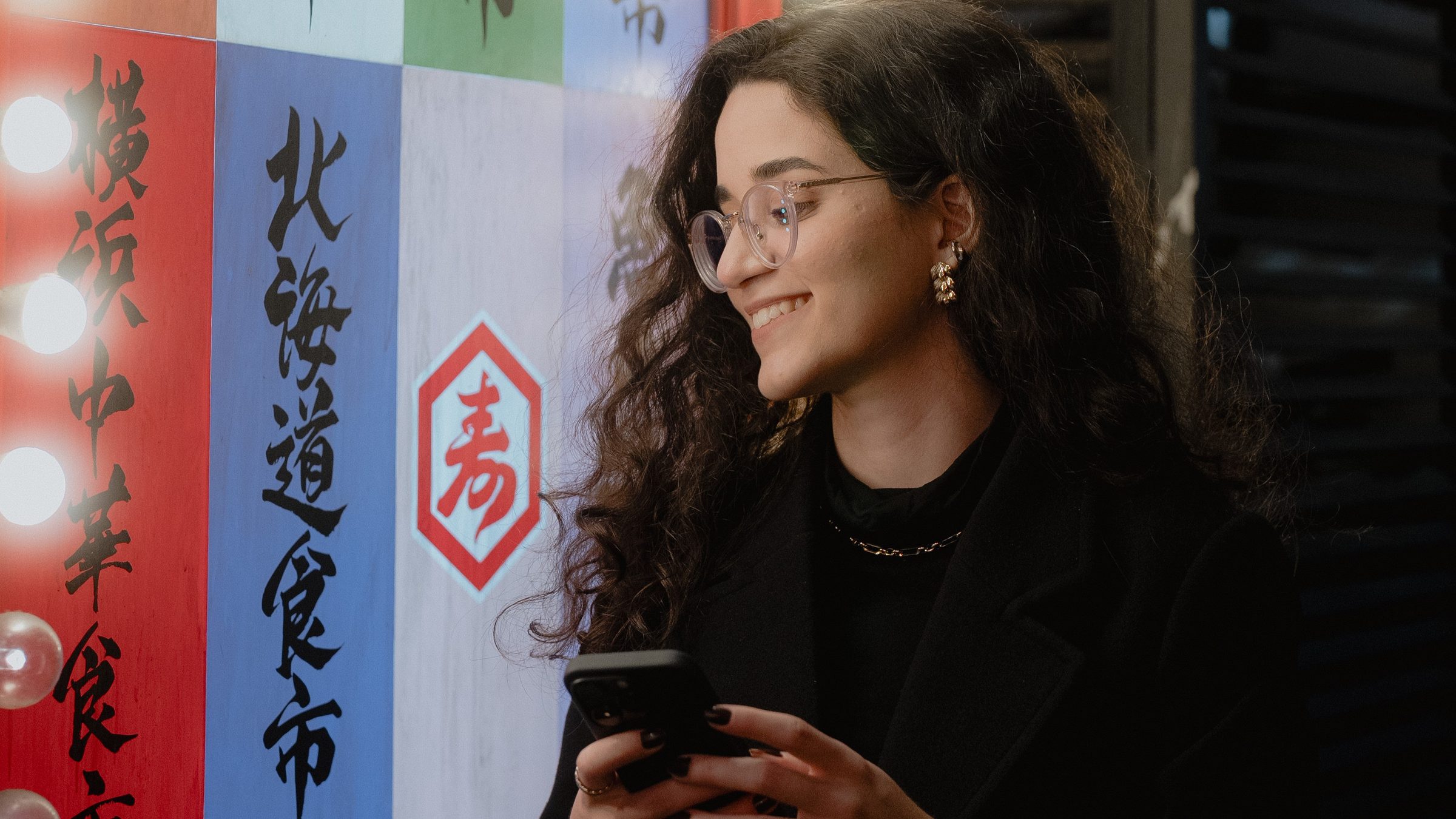 Mulher sorrindo ao olhar o celular, em frente a um painel colorido com caligrafia japonesa